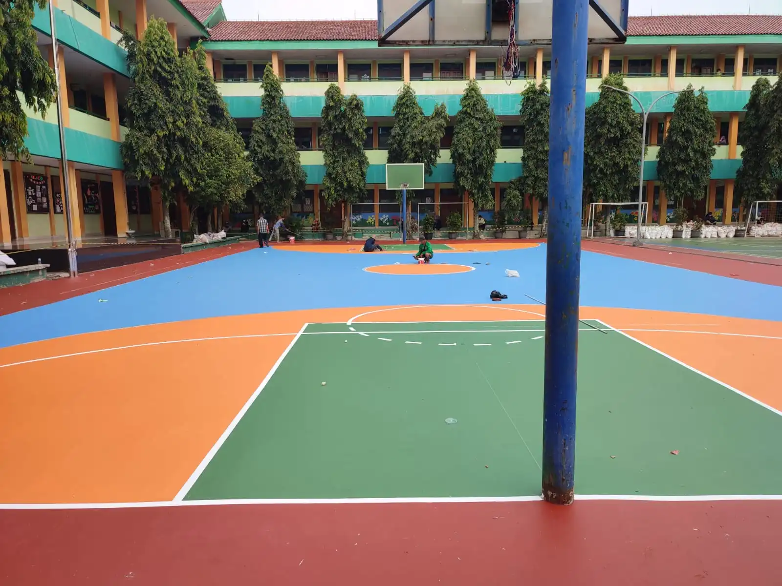 Lapangan basket dengan cat berwarna hijau, oranye, dan biru yang sedang dikerjakan